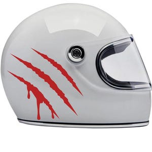 Op de afbeelding: Witte motorhelm met een helder vizier en rode klauwmarkeringen. De helm heeft een glanzende afwerking en een klassiek ontwerp, geschikt voor motorrijders. Hij heeft een helder vizier en een kinband.