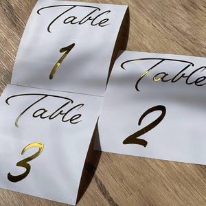 May include: White table number cards with gold foil lettering. Each card has the word "Table" above a number. Numbers 1, 2, and 3 are visible. The cards are on a light brown wooden surface.