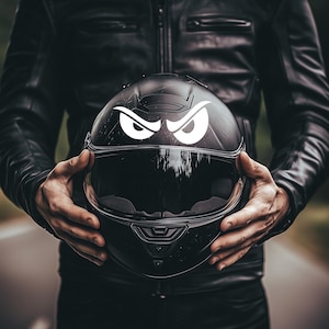 Pode incluir: Capacete de motocicleta preto com viseira transparente e um gráfico branco de olhos zangados. O capacete é segurado por uma pessoa vestindo uma jaqueta de couro preta, com uma estrada desfocada ao fundo.