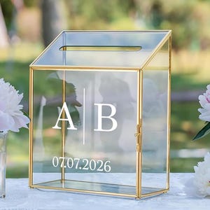 Peut inclure: Une boîte à cartes en verre transparent avec un cadre en métal doré. La boîte a un dessus incliné avec une fente et une porte à charnière. Les initiales blanches "AB" et la date "07.07.2026" sont imprimées sur le devant. L'arrière-plan est flou avec de la verdure et des fleurs.