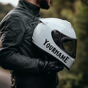 Può includere: Casco da moto bianco con visiera nera e la scritta "YOURNAME" in lettere nere. Il casco è tenuto da una persona che indossa una giacca e guanti di pelle neri. Lo sfondo è sfocato.