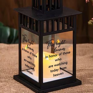 Op de afbeelding: Zwarte metalen lantaarn met glazen panelen, met de tekst "This light burns in honor of those who are watching today from heaven." De lantaarn bevat een brandende kaars. Een decoratief of herdenkingsitem.