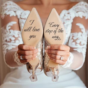 Può includere: Un paio di scarpe con tacco alto dorate e glitterate con tacchi trasparenti. Le suole riportano le frasi "I will love you" e "Every step of the way" scritte in nero. La persona indossa un abito bianco.