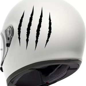 Peut inclure: Casque de moto blanc avec une visière transparente. Le casque présente quatre autocollants noirs en forme de griffes sur le côté. Le rembourrage intérieur est noir et gris.