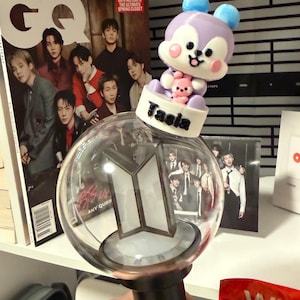 May include: A clear, spherical light stick with a black handle, featuring a white and silver logo. A purple and white cartoon figure with blue ears and a pink bear sits atop the light stick. The name "Tasia" is printed on the base.