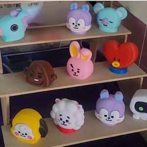 May include: A wooden display shelf holds a collection of colourful character figurines. The figurines include a blue koala, a purple bunny, a red heart, and a yellow character. The shelf is made of light-coloured wood.