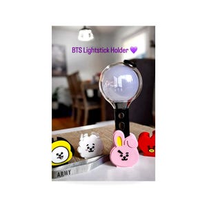 May include: A BTS lightstick holder with a purple heart graphic. The image shows a lightstick with a clear orb and black handle, along with character-shaped holders in yellow, white, pink, and red. The word "ARMY" is visible on a strap.