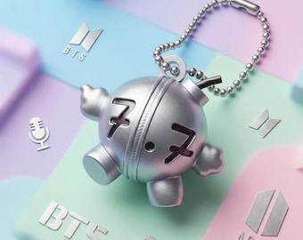 May include: A silver-colored spherical keychain with a ball chain, featuring the number 7 embossed on the front. The object has a small protruding fuse and four rounded feet. The design includes the text BTS and ARMY with microphone icons.