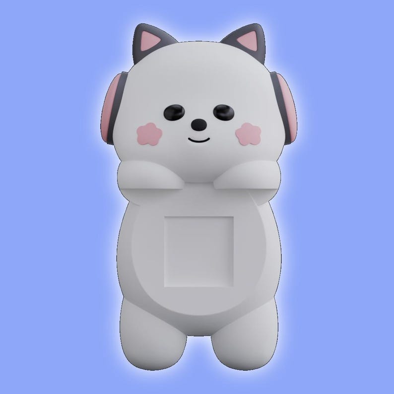 May include: A white cat-shaped phone stand featuring dark gray ears with pink inner details, pink cheek accents, black eyes and nose, and pink headphones on the sides. The center of the body has a square cutout for holding a mobile device.