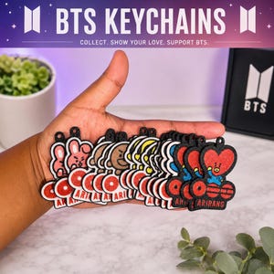 Peut inclure: Plusieurs porte-clés BTS avec des personnages variés, tenus dans une main. Chaque porte-clé présente un cercle rouge avec le texte "ARIRANG" ou "AR" en blanc. La bannière supérieure indique "BTS KEYCHAINS".