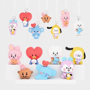 Puede incluir: Una colección de llaveros de personajes coloridos. Los llaveros presentan varias figuras de dibujos animados en tonos de rosa, azul, amarillo y marrón, cada uno sosteniendo un personaje más pequeño a juego. Están unidos a cadenas plateadas.