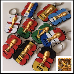 Puede incluir: Una colección de llaveros con el trofeo de la Copa Mundial de la FIFA. Cada llavero presenta un trofeo dorado sobre un diseño de bandera, incluyendo banderas de Inglaterra, Francia, Brasil y Alemania. La palabra "FIFA" está impresa en cada llavero.
