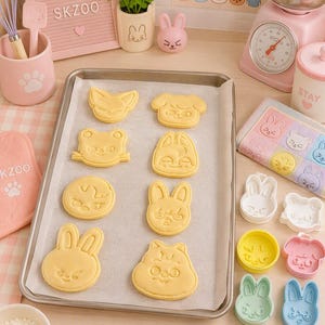 Peut inclure: Une plaque de cuisson rectangulaire en métal recouverte de papier sulfurisé contient huit biscuits jaune clair en forme d'animaux. À côté se trouvent des emporte-pièces en plastique colorés, un rouleau à pâtisserie en bois avec le texte STAY.