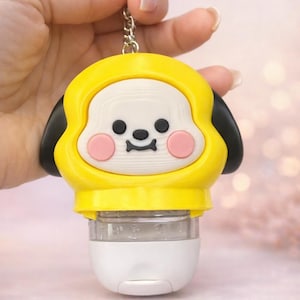 May include: A yellow and white character-shaped hand sanitizer holder with black ears, pink cheeks, and a small black nose and mouth. The holder features a silver metal keychain ring at the top and a clear plastic bottle with a white cap at the bottom.