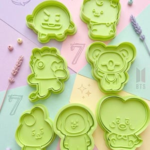 Peut inclure: Un ensemble de sept emporte-pièces en plastique vert clair représentant diverses formes de personnages de dessins animés. Ils sont disposés sur un fond multicolore avec des illustrations de baleines, le chiffre 7, des étoiles et le texte AESTHETIC BT21 x BTS BAKING.