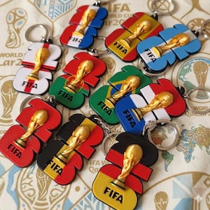 Könnte beinhalten: Eine Sammlung von FIFA-Weltmeisterschafts-Schlüsselanhängern mit dem goldenen Pokalsymbol, das auf verschiedenen Nationalflaggen-Designs zentriert ist. Jeder Schlüsselanhänger hat einen Metallring und zeigt den Schriftzug FIFA in Schwarz.