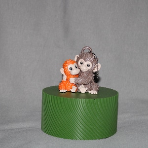 Peut inclure: Une figurine de deux singes enlacés, un orange et un gris, est posée sur une base verte cylindrique. Le singe gris a un porte-clés attaché. La base présente un motif rainuré texturé.
