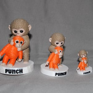 Op de afbeelding: Drie apenbeeldjes. Elk beeldje toont een grotere bruine aap die een kleinere oranje aap omhelst. De beeldjes staan op witte sokkels met het woord "PUNCH" erop gedrukt. De maten variëren.