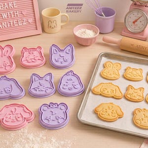 Peut inclure: Un ensemble d'emporte-pièces en plastique rose et violet en forme de visages d'animaux, disposés à côté d'une plaque de cuisson en métal contenant huit biscuits cuits aux motifs assortis, un rouleau à pâtisserie et un tableau à lettres rose.