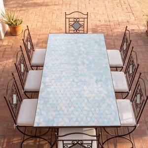 May include: An outdoor dining set featuring a rectangular table with a mosaic tile top in shades of blue and white. The set includes eight wrought iron chairs with white cushions, arranged on a brick patio. The table is approximately 72 inches long.