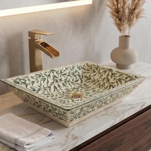 May include: A square ceramic bathroom sink with a green and white leaf pattern. A gold tap and drain are visible. A black soap dispenser, folded white towel, and a vase with dried plants are on the countertop.