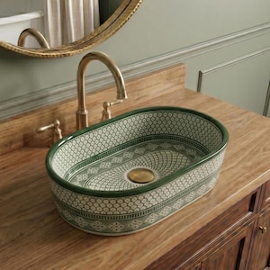 May include: An oval ceramic bathroom sink with a green and white geometric pattern. The sink is set on a wooden vanity with a gold tap and drain. A gold-framed mirror hangs above the sink.