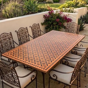 Puede incluir: Una mesa de comedor rectangular para exteriores con una superficie de tono cobrizo que presenta un patrón geométrico repetitivo, rodeada por ocho sillas de metal con respaldos de volutas intrincadas y cojines claros sobre un patio de piedra.