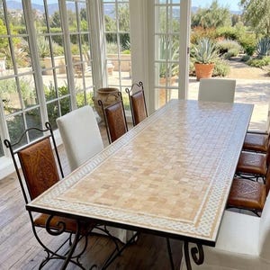 Può includere: Un lungo tavolo da pranzo rettangolare con piano a mosaico e bordo decorativo. Il tavolo è circondato da otto sedie, alcune con sedili in pelle e altre con coperture bianche. Le gambe in metallo del tavolo hanno un design a volute.