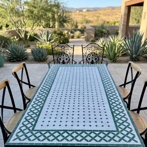 Op de afbeelding: Eetset voor buiten met een rechthoekige tafel met een wit en groen betegeld blad en een zwart metalen frame. Vier stoelen met houten rugleuningen en geweven zittingen omringen de tafel. De achtergrond toont een woestijnlandschap.