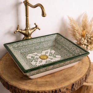 Op de afbeelding: Een rechthoekige keramische wastafel met een groen en wit geometrisch patroon. De wastafel staat op een houten oppervlak, met een vintage messing kraan op de achtergrond. De wastafel heeft een messing afvoer en een groene rand.