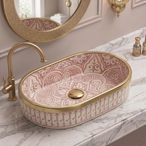 May include: Oval ceramic bathroom sink with a pink and white floral design, gold trim, and a gold tap. The sink is set on a marble countertop, with a gold-framed mirror and wall sconces in the background.