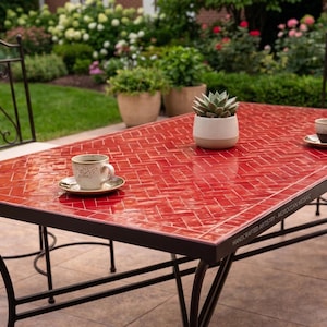 Puede incluir: Una mesa rectangular para exteriores con una tapa de mosaico rojo y un marco de metal negro. La mesa tiene una suculenta en una maceta blanca y dos tazas de café. La mesa está rodeada de sillas y vegetación.