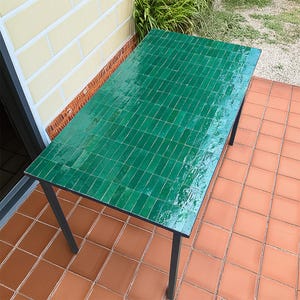 以下が含まれることがあります： 鮮やかな緑色のタイル張りの天板と黒い金属製の脚が付いた長方形のテーブル。テーブルの表面は、小さな長方形のタイルで構成されており、質感のある外観を作り出しています。テーブルの長さは約152cm、幅は約76cmです。