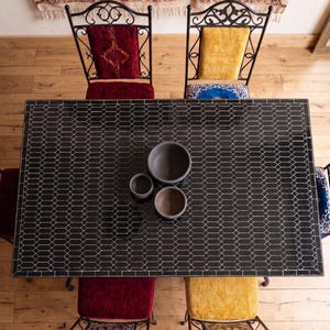 Op de afbeelding: Bovenaanzicht van een eettafel met een zwart geometrisch betegeld blad. De tafel wordt omringd door smeedijzeren stoelen met rode en gele fluwelen kussens. Drie grijze keramische kommen staan op de tafel.