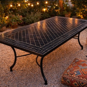 Può includere: Un tavolo rettangolare da esterno con piano piastrellato nero e gambe in metallo nero curve. Il tavolo è circondato da cuscini decorativi e si trova su una superficie di ghiaia. Le luci della stringa sono visibili sullo sfondo.