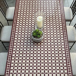 Könnte beinhalten: Ein rechteckiger Esstisch für den Außenbereich mit einer Mosaik-Tischplatte in einem geometrischen Muster aus Burgunderrot und Creme. Ein Glaskerzenhalter mit einer brennenden Kerze und eine kleine Topf-Sukkulente befinden sich auf dem Tisch. Vier Metallstühle mit gepolsterten Sitzen umgeben den Tisch.