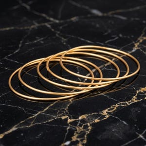May include: A set of five thin, circular gold-toned bangles arranged in an overlapping formation. Each bangle features a smooth, polished surface with a consistent round profile, creating a stackable jewelry set with a metallic finish.