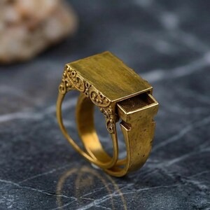 May include: A gold-colored ring with a rectangular top and a small drawer that pulls out. The ring has ornate filigree detailing on the sides of the rectangular top. The ring is made of a shiny, metallic material.