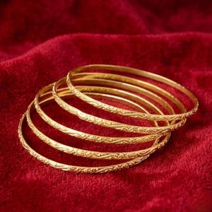 May include: Six gold bangles with detailed floral designs rest on a deep red velvet surface. The thin, circular bangles reflect light. The background is a rich, textured red.