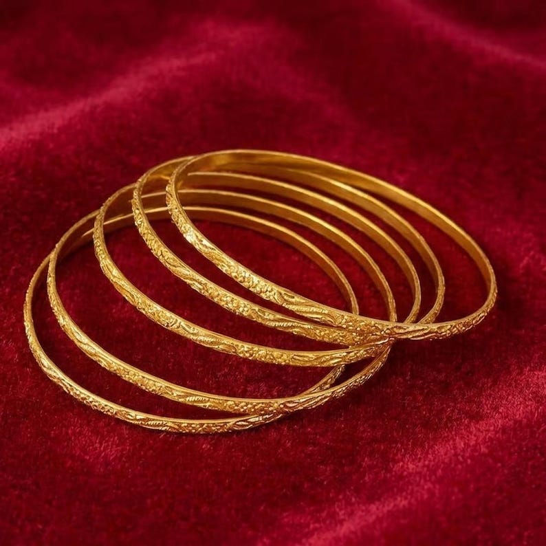 May include: A set of gold bangles with detailed floral designs. The bangles are arranged on a deep red velvet surface, creating a luxurious display. The gold colour of the bangles contrasts with the rich red background, creating a striking visual.