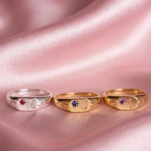 Könnte beinhalten: Drei Siegelringe mit eingravierten Buchstaben und kleinen eingesetzten Edelsteinen. Der linke Ring ist silberfarben mit einem roten Stein und dem Buchstaben M. Der mittlere Ring ist goldfarben mit einem blauen Stein und dem Buchstaben S. Der rechte Ring ist goldfarben mit einem violetten Stein und dem Buchstaben J.