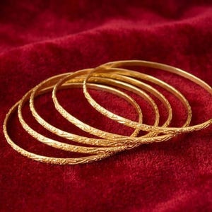 May include: Six gold bangles with detailed floral designs, displayed on a deep red velvet background. The bangles' golden colour is accentuated by the red backdrop. These bangles are a traditional jewellery piece.