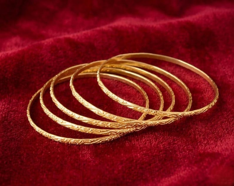 May include: Six gold bangles with detailed floral designs, displayed on a deep red velvet background. The bangles' golden colour is accentuated by the red backdrop. These bangles are a traditional jewellery piece.