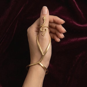 Puede incluir: Un adorno de mano dorado que presenta un anillo con forma de serpiente conectado por una barra de metal delgada a un brazalete. El anillo de serpiente envuelve el pulgar, mientras que la estructura de metal lisa en forma de Y baja hasta la muñeca.