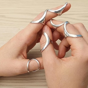 May include: A collection of silver-toned rings in various designs. The rings are worn on multiple fingers, showcasing a range of shapes, including curved and pointed styles. The jewelry is displayed on a light-colored surface.