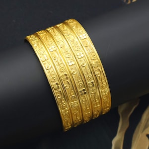 Puede incluir: Un conjunto de cinco brazaletes dorados con un patrón repetitivo de flores y enredaderas en relieve. Cada brazalete tiene un ancho constante y una superficie texturizada con detalles realzados, creando una apariencia uniforme en todo el conjunto.