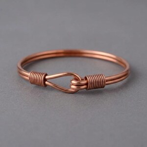 Puede incluir: Una pulsera de alambre de cobre con un diseño de doble hebra que forma un cierre de gancho. Los extremos del alambre están enrollados firmemente alrededor de la banda principal para asegurar el mecanismo de bucle y gancho, creando una forma circular.