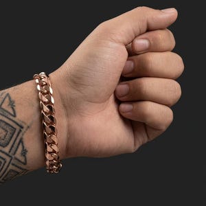 May include: A rose gold-toned cuban link chain bracelet featuring thick, interlocking, polished metal links that create a continuous, heavy-duty design. The bracelet is worn around the wrist, showcasing a reflective, metallic finish throughout.