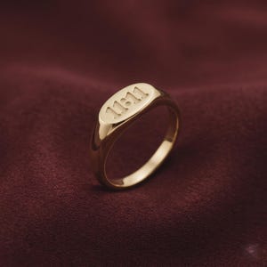 May include: A gold-colored signet ring with the engraved number "11:11" on the oval face. The ring is set against a dark red background, highlighting its polished finish. A classic piece of jewelry.