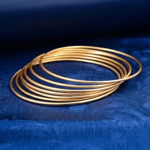 Peut inclure: Une pile de bracelets fins de couleur or. Les bracelets sont circulaires et semblent être en métal. Ils sont disposés sur une surface bleu foncé texturée.